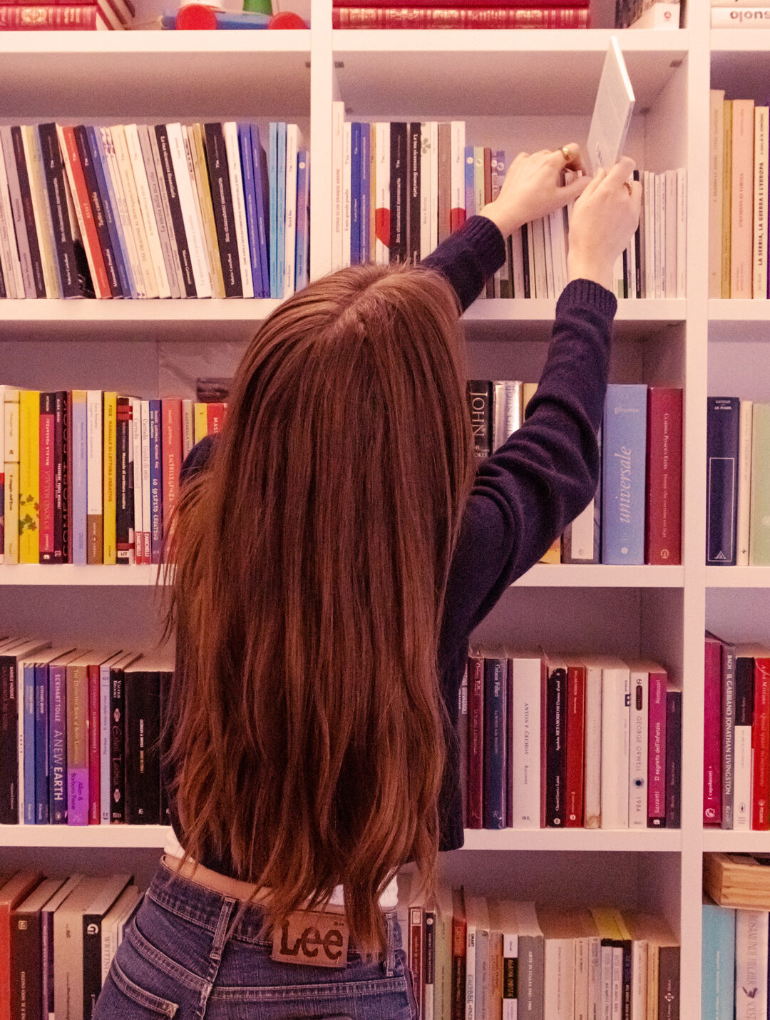 alt="ragazza di spalle prende un libro da una libreria"
